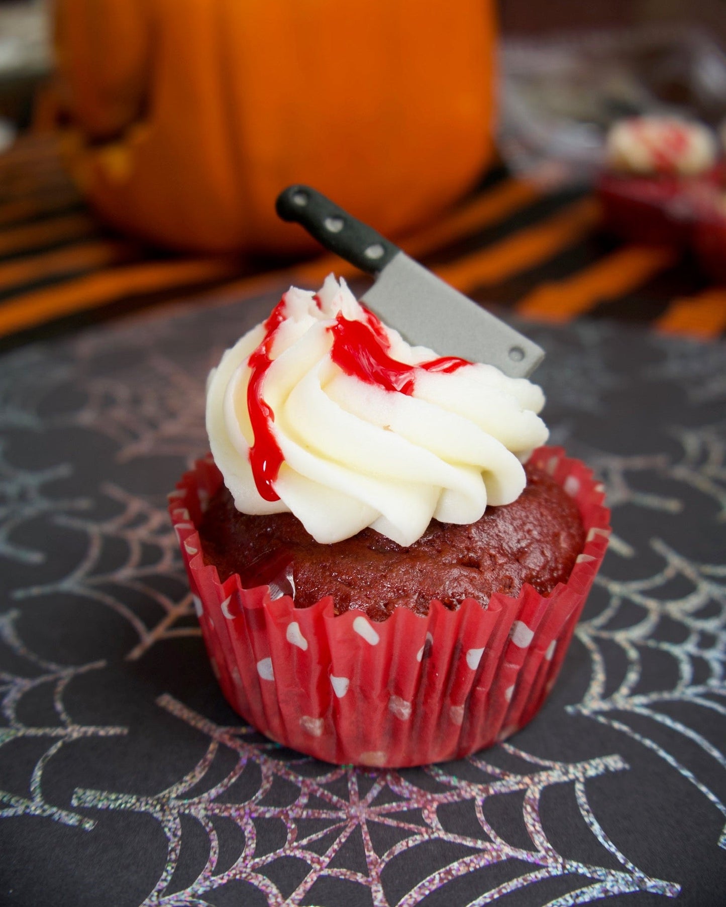 Red velvet cupcake with white frosting and red sauce, stabbed with a knife, on a Halloween-themed tablecloth.