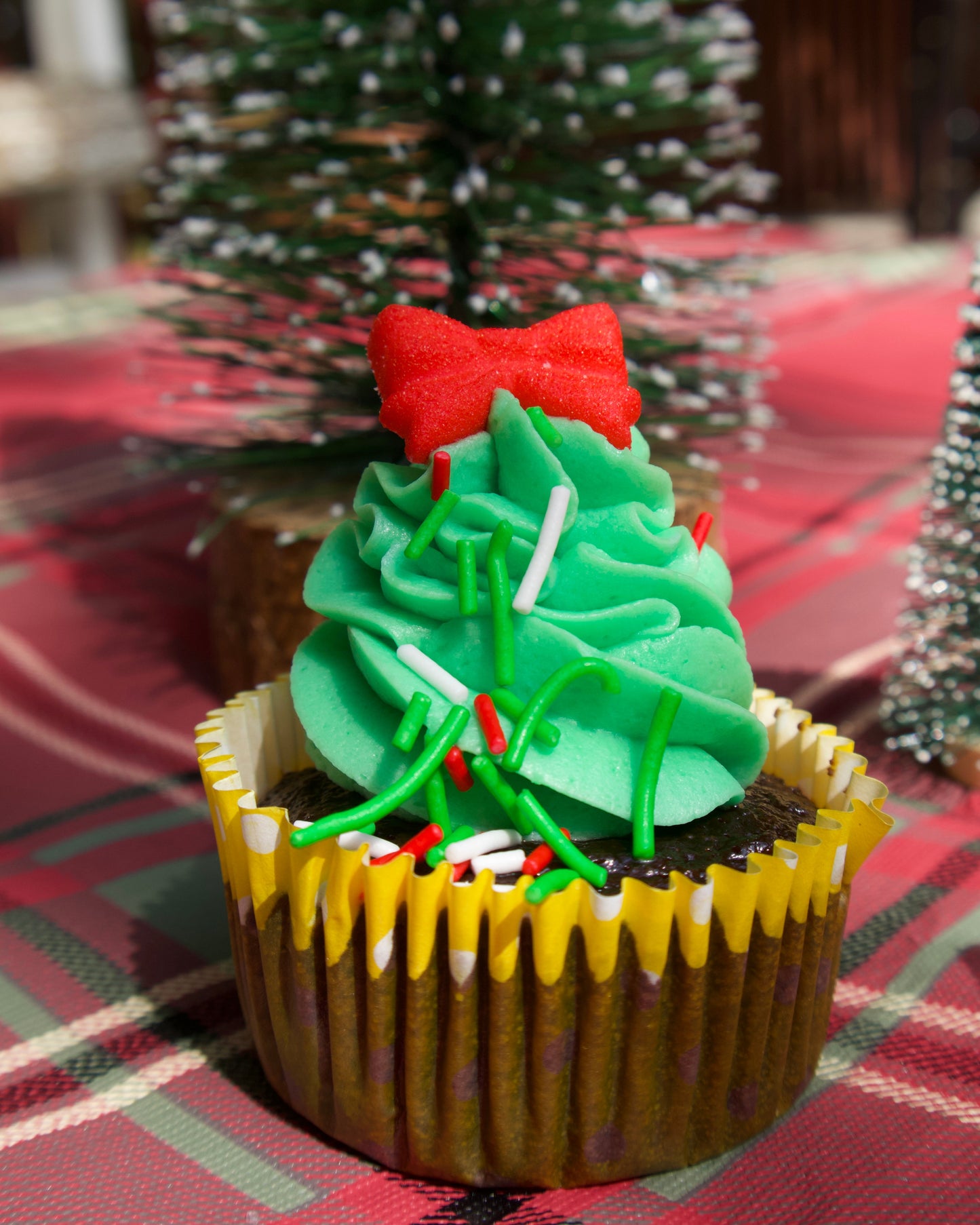 Cupcake decorated like a Christmas tree with a red bow on a plaid tablecloth.